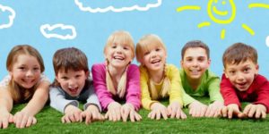 Троє хлопчиків і троє дівчат лежать на траві на фоні намальованого неба з усміхненим сонцем і хмаринками. Діти радісні, усміхнені